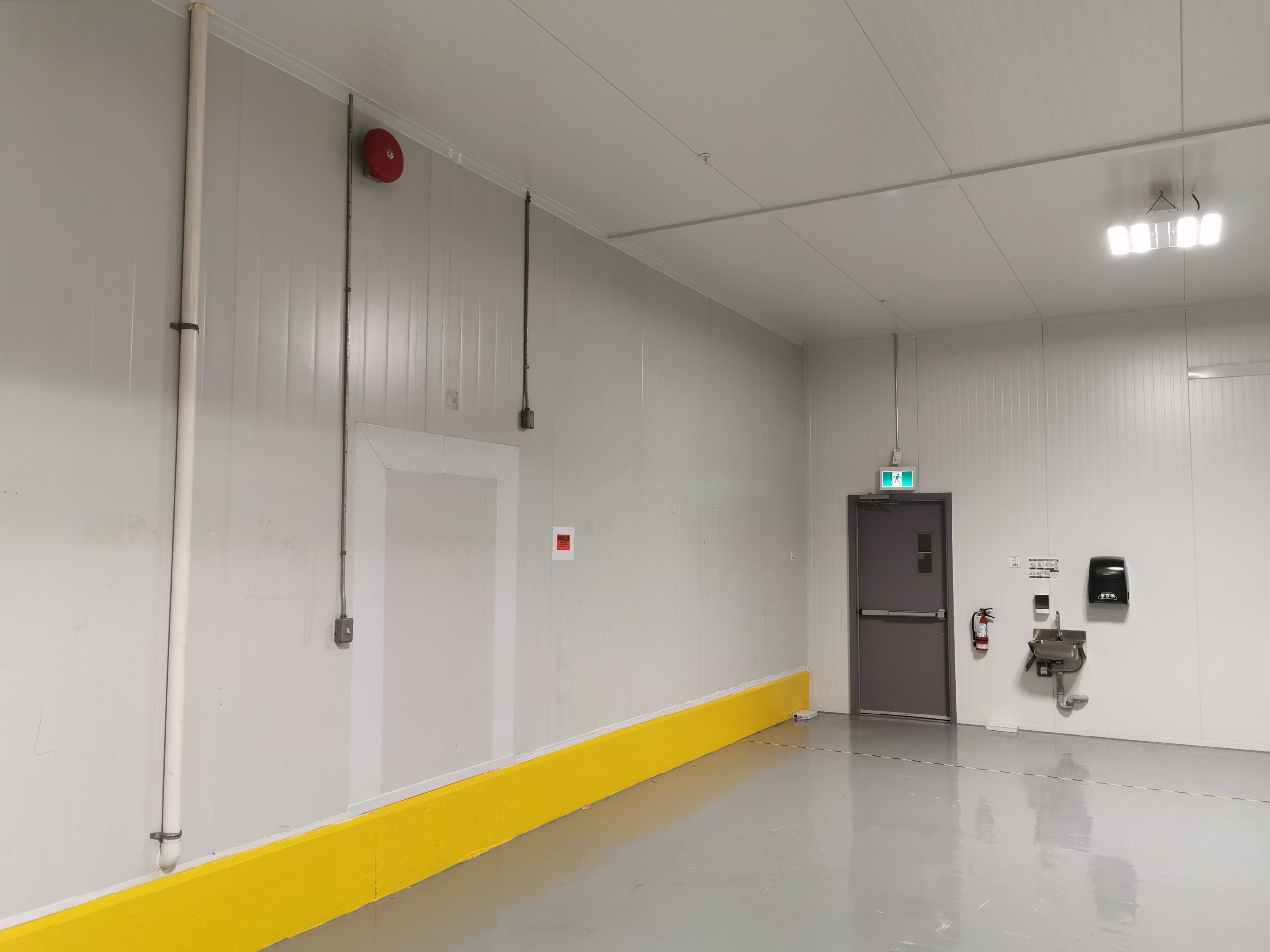 Industrial room with white walls, yellow floor bumper, grey exit door, fire alarm, sink, and emergency signage.